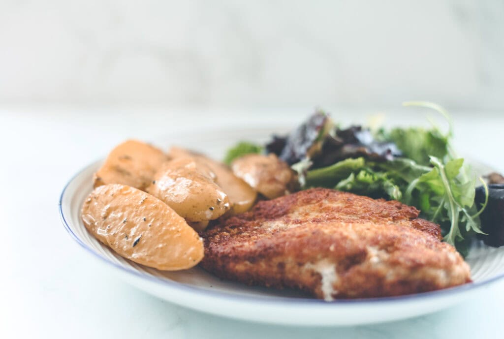 A long shot of a dish of chicken parmesan with potatoes and salad.